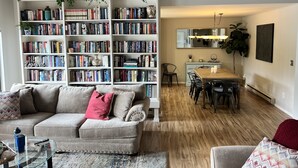 A cozy living room with a sofa, a coffee table, and a bookshelf filled with books. There is a dining area with a table and chairs.