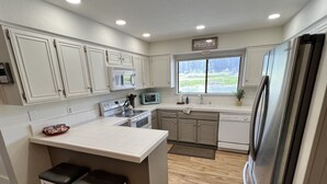 A modern kitchen with white cabinetry, a central island with stools, stainless steel appliances, and a view of greenery outside.