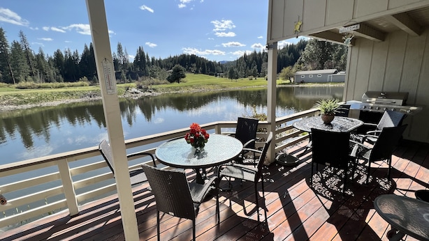 A covered outdoor deck with a table and chairs, overlooking a lake and forested area.