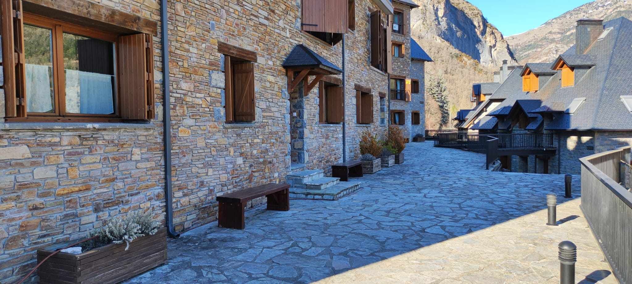 Un bâtiment en pierre avec des volets en bois, une cour pavée de pierres et une chaîne de montagnes en arrière-plan.