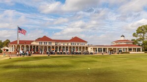 Un edificio grande e histórico con tejado rojo y múltiples columnas, rodeado por un césped cuidado y un campo de golf.