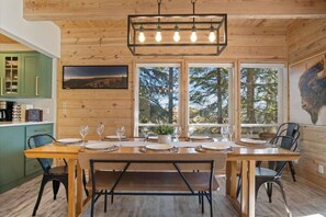 A wooden interior with a dining table set for four, a kitchen with green cabinets, and a large window with a forest view.