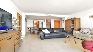 Una sala de estar con televisor de pantalla plana, un sofá de cuero negro, un sofá color beige, una mesa de centro de vidrio y armarios de madera.