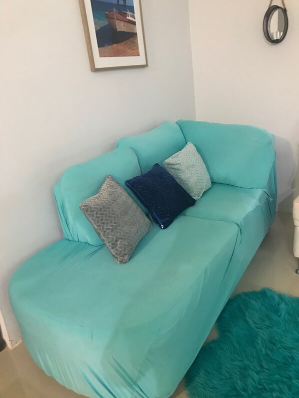 A teal sofa with three cushions in a room with a framed picture on the wall and a round mirror on the right.