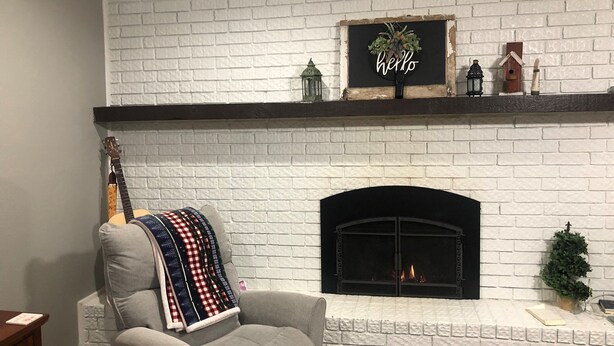 Un salón con chimenea de ladrillo blanco, una guitarra colgada, un sillón gris y un letrero que dice 'hello' sobre la repisa.