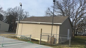 Un pequeño edificio con techo inclinado, una cancha de tenis cercada y dos farolas.