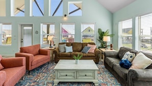 A spacious living room with a brown sofa, two armchairs, a coffee table, and a patterned rug. Large windows with blinds provide natural light and views of the outside. - ☀ Tranquil beach! 100ft to Beach ~ Ocean Views, Pets  (Galveston)
