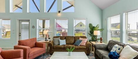 Een ruime woonkamer met een bruine bank, twee fauteuils, een salontafel en een kamerkleed met een patroon. Grote ramen met zonwering laten natuurlijk licht en uitzicht naar buiten toe.