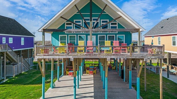 A multi-level wooden structure with a balcony, surrounded by a grassy area and adjacent buildings. - ☀ Tranquil beach! 100ft to Beach ~ Ocean Views, Pets  (Galveston)