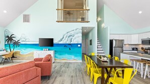 A dining area with a mural of a beach scene, a TV, and a kitchen with white cabinets and a stainless steel refrigerator. - ☀ Tranquil beach! 100ft to Beach ~ Ocean Views, Pets  (Galveston)