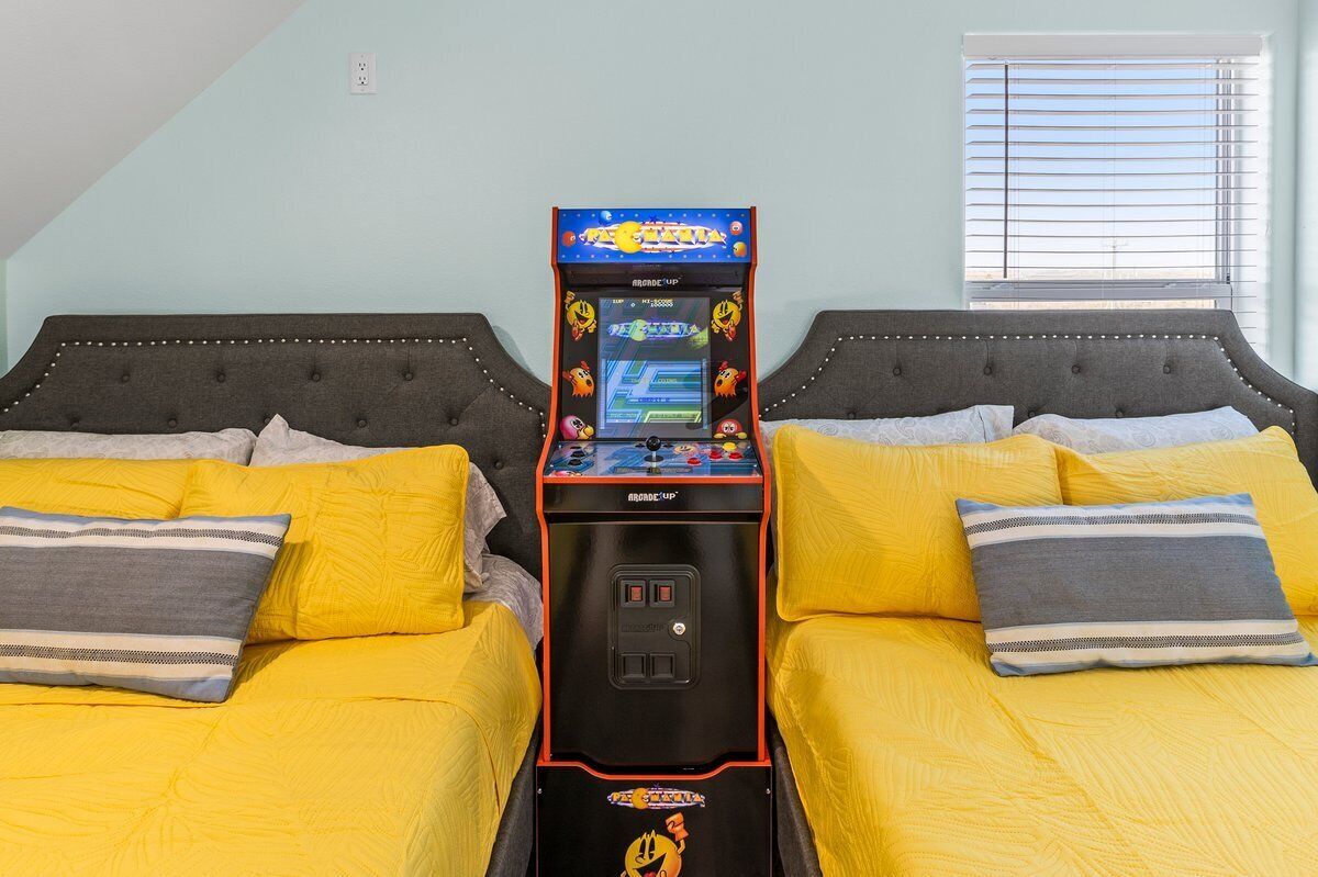 Una habitación con dos camas, cada una con ropa de cama amarilla y almohadas grises, y una máquina recreativa retro en el centro.