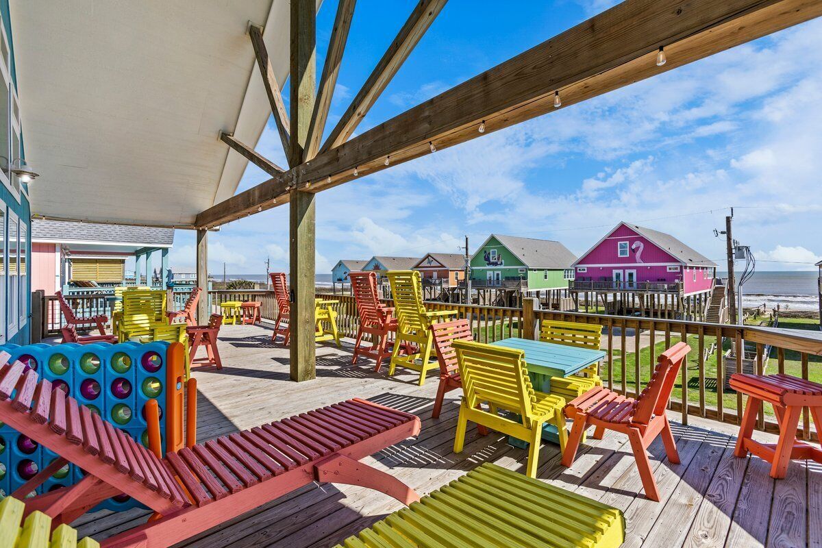 A covered outdoor deck with colorful chairs and tables, overlooking a beach and colorful buildings.