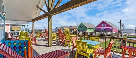 A covered outdoor deck with colorful chairs and tables, overlooking a beach and colorful buildings.