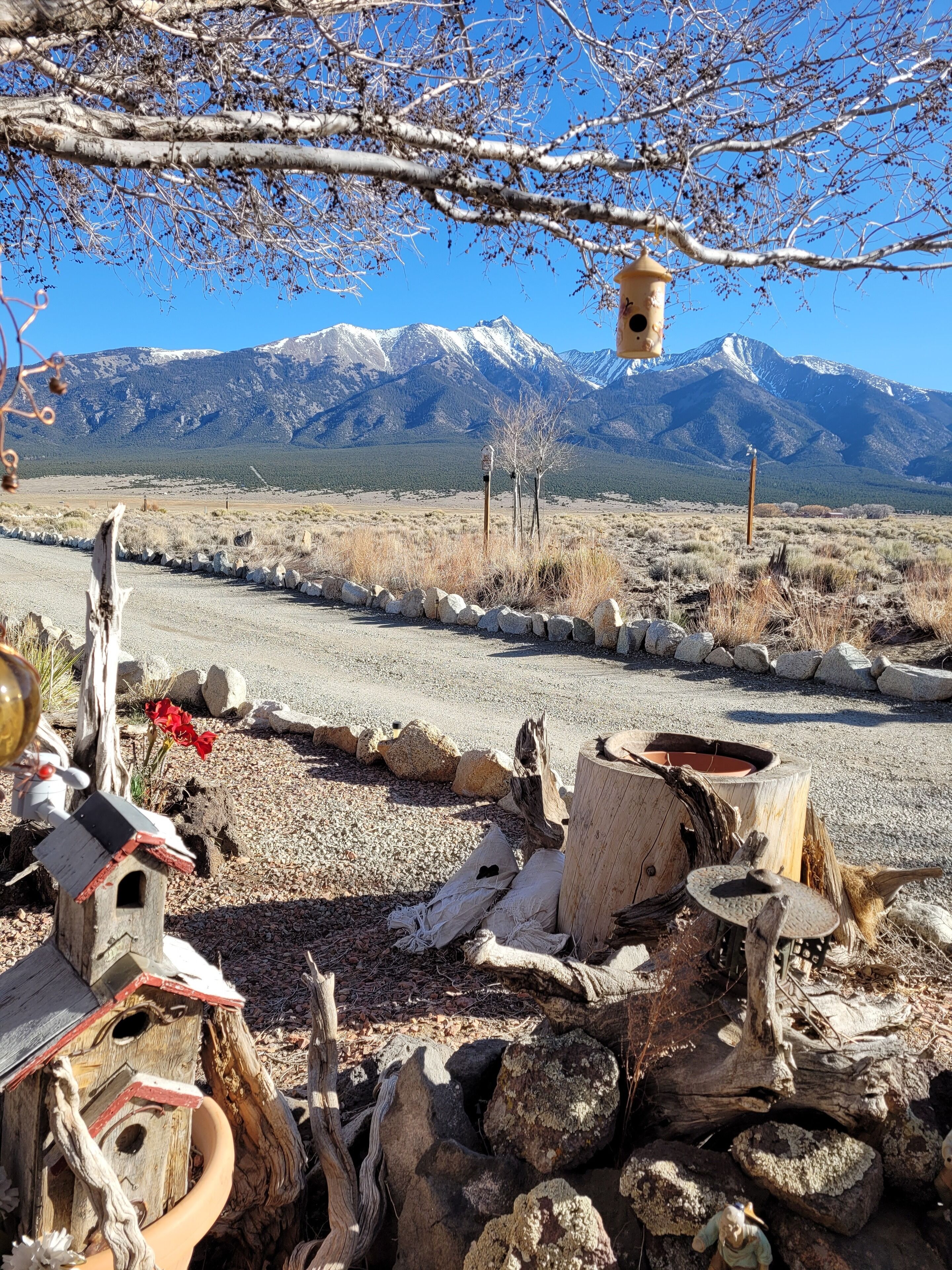 A rustic outdoor setting with a wooden birdhouse, a rustic fire pit, and a clear view of snow-capped mountains in the background.