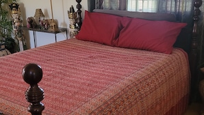 Cama bem arrumada com colcha estampada, cabeceira de madeira e travesseiro vermelho. Pequena mesa com abajur e objetos decorativos ao lado da cama.
