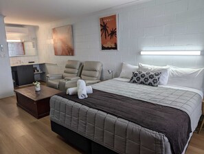 Habitación de hotel con cama, dos sillones reclinables, una mesita, una mesita de noche, una lámpara, un cuadro y una zona de cocina.