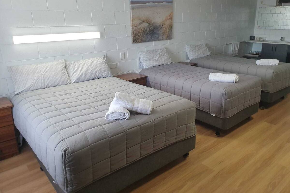 Ein Hotelzimmer mit drei Betten, jedes mit einer gesteppten Bettdecke und Kissen. Darüber hängt ein Gemälde an der Wand, darunter ein kleiner Tisch mit einer Lampe, und der Boden ist mit Holz versehen.