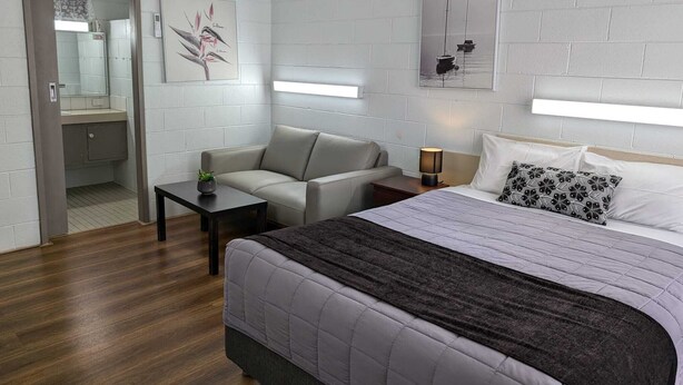 A modern hotel room with a bed, a sofa, a small table, and a nightstand. There is a mirror, a painting, and a window with blinds. - Morwell Parkside Motel (Morwell)