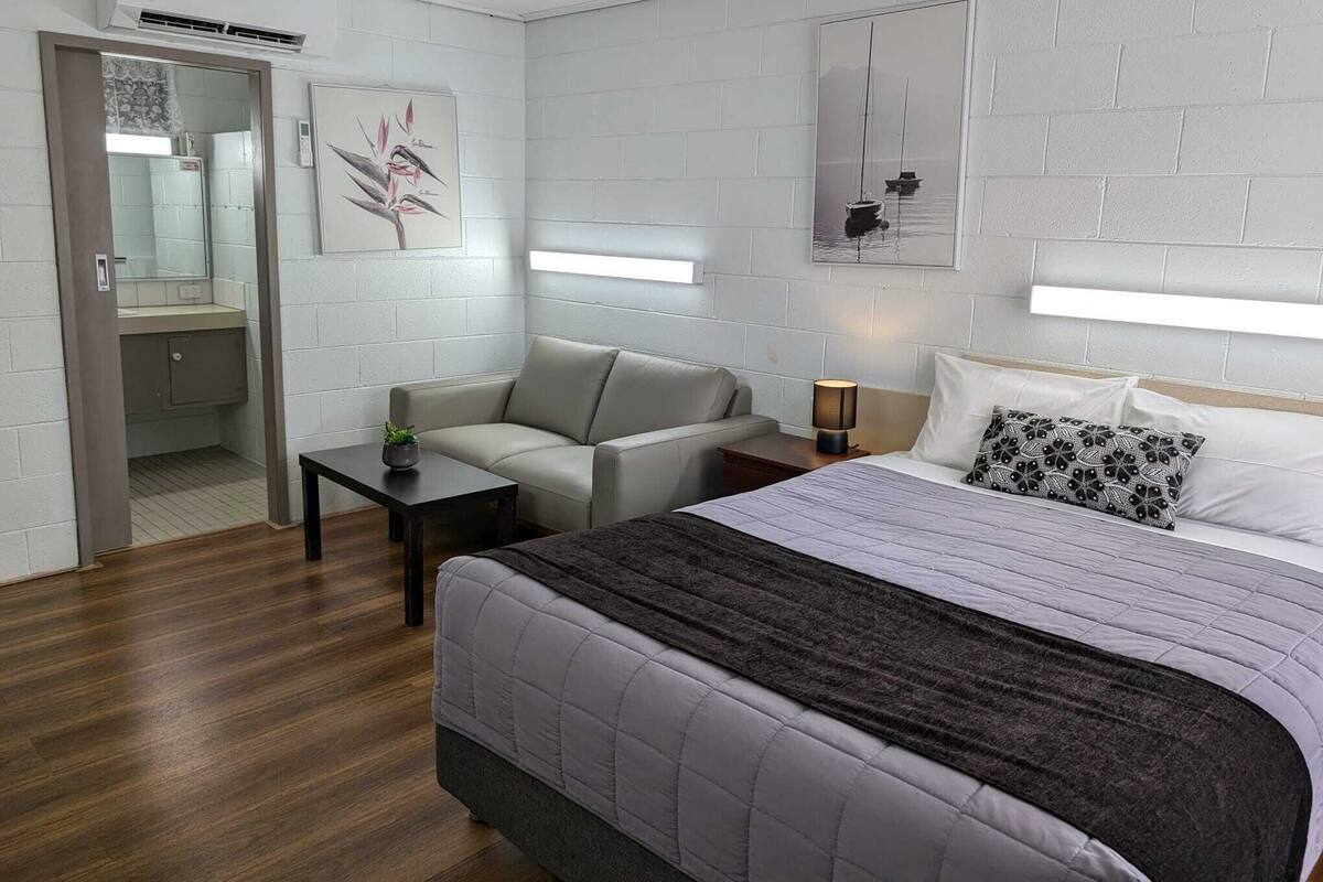 Ein modernes Hotelzimmer mit einem Bett, einem Sofa, einem kleinen Tisch und einem Nachttisch. Es gibt einen Spiegel, ein Gemälde und ein Fenster mit Jalousien.