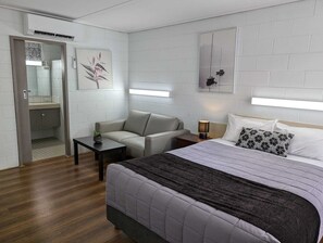 A modern hotel room with a bed, a sofa, a small table, and a nightstand. There is a mirror, a painting, and a window with blinds. - Morwell Parkside Motel (Morwell)