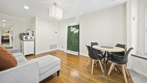 Ett modernt vardagsrum med en grå soffa, ett matbord med fyra stolar, en köksdel med mikrovågsugn och en grön vägg med en neonreklamskylt.