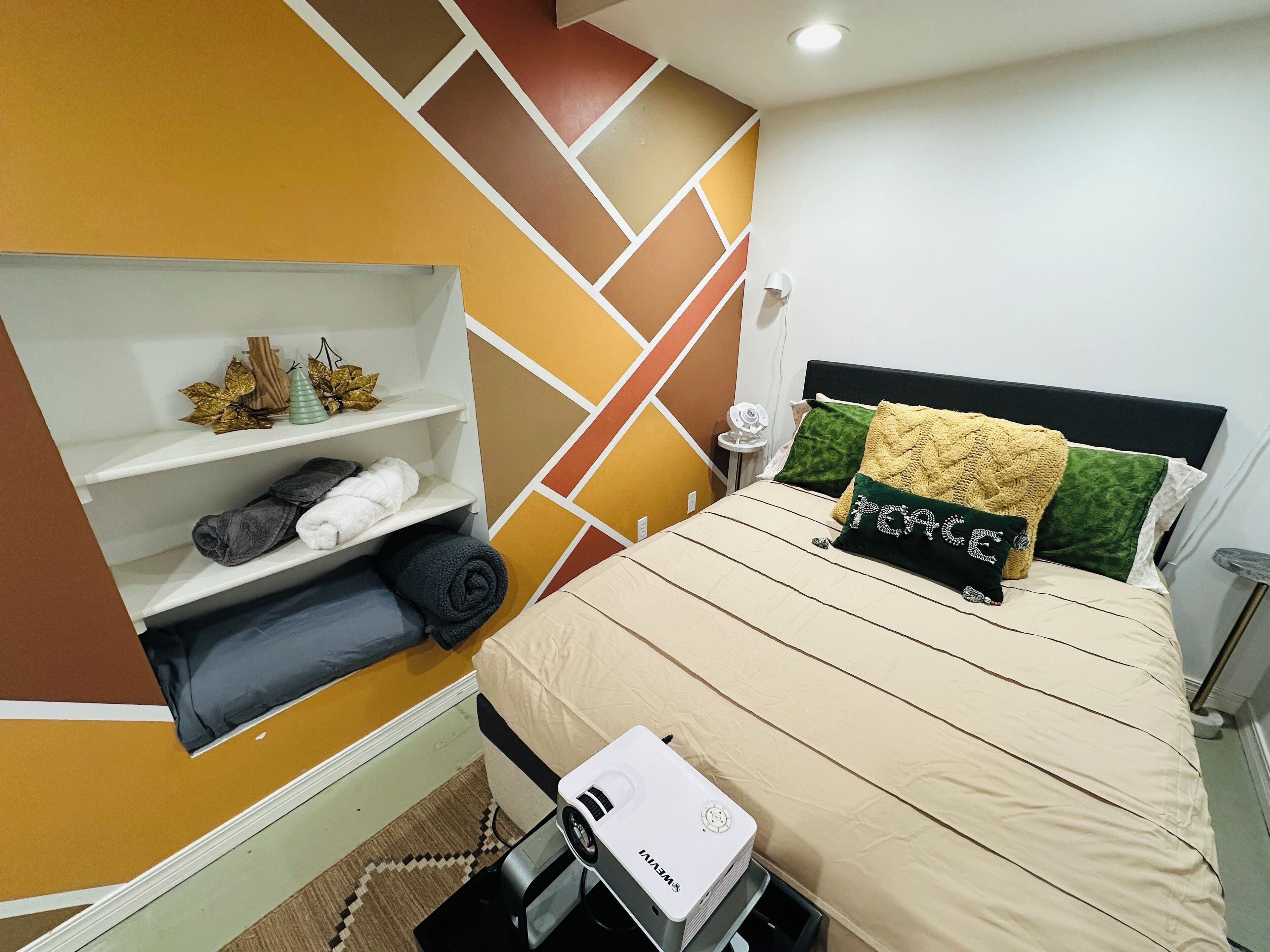 A bedroom with a bed, a shelf with towels, a projector, and a wall with geometric patterns.