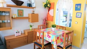 A dining area with a table covered in a floral tablecloth, wooden chairs, a shelf with kitchen items, and a window with a view.