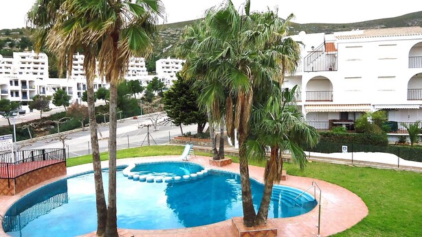 A swimming pool with palm trees, a slide, and a white building with balconies in the background.