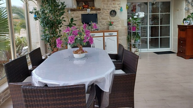 Een overdekt terras met een ronde tafel en rieten stoelen, omgeven door potplanten en een stenen muur.