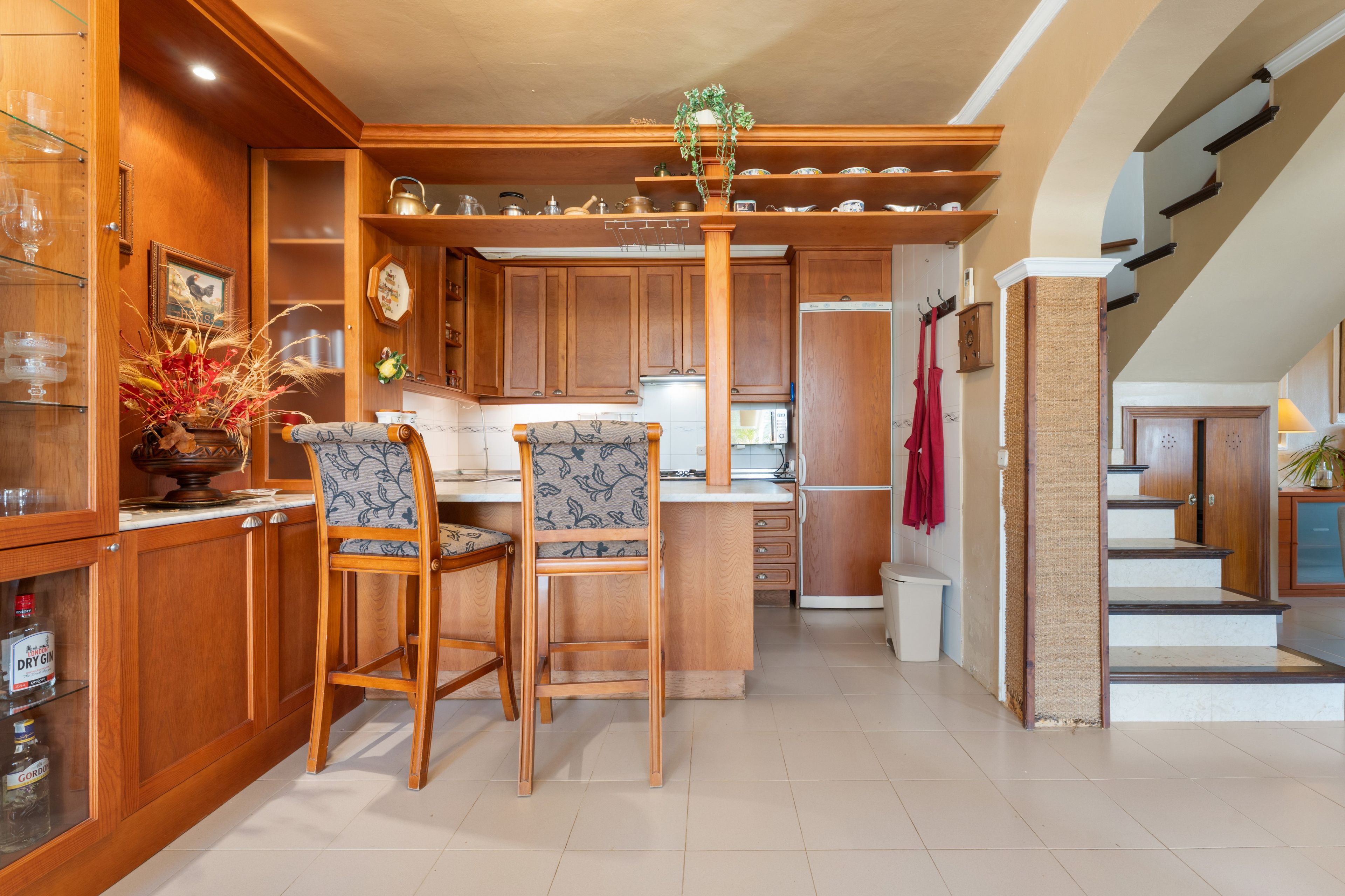 Una cocina con armarios de madera, una isla central, dos taburetes de bar y una planta colgante.