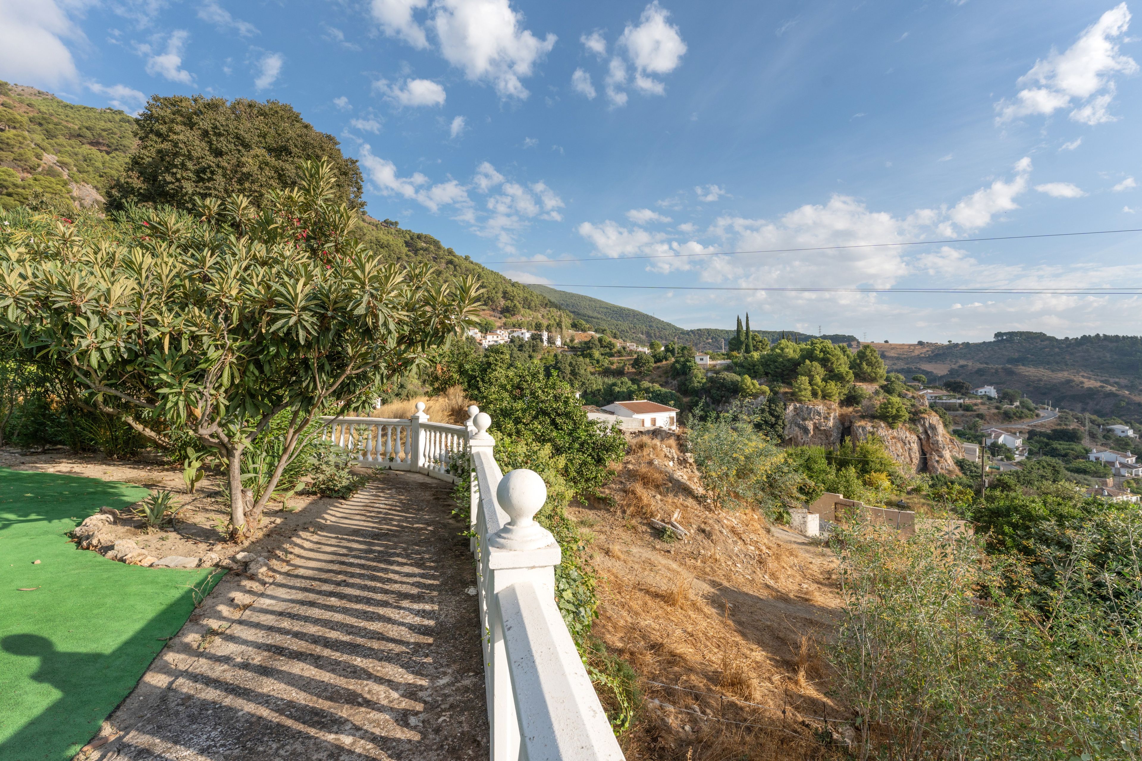 Un sendero lleva a una ladera con baranda blanca, rodeada de vegetación y con vistas a un pueblo.