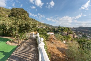 A pathway leading to a hillside with a white railing, surrounded by greenery and overlooking a village.