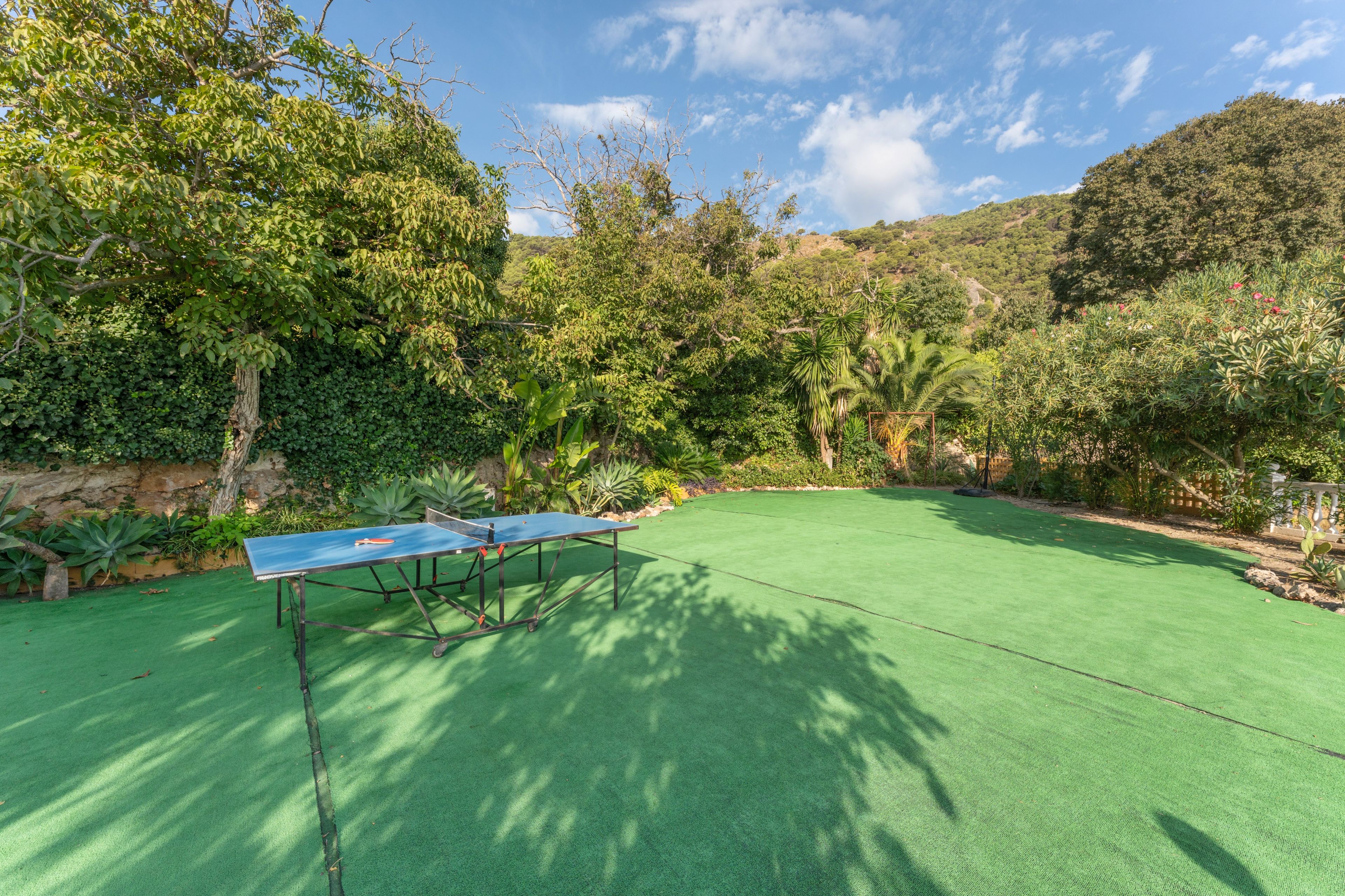 Una mesa de ping pong montada sobre césped artificial verde en un jardín exuberante con diversas plantas y árboles.