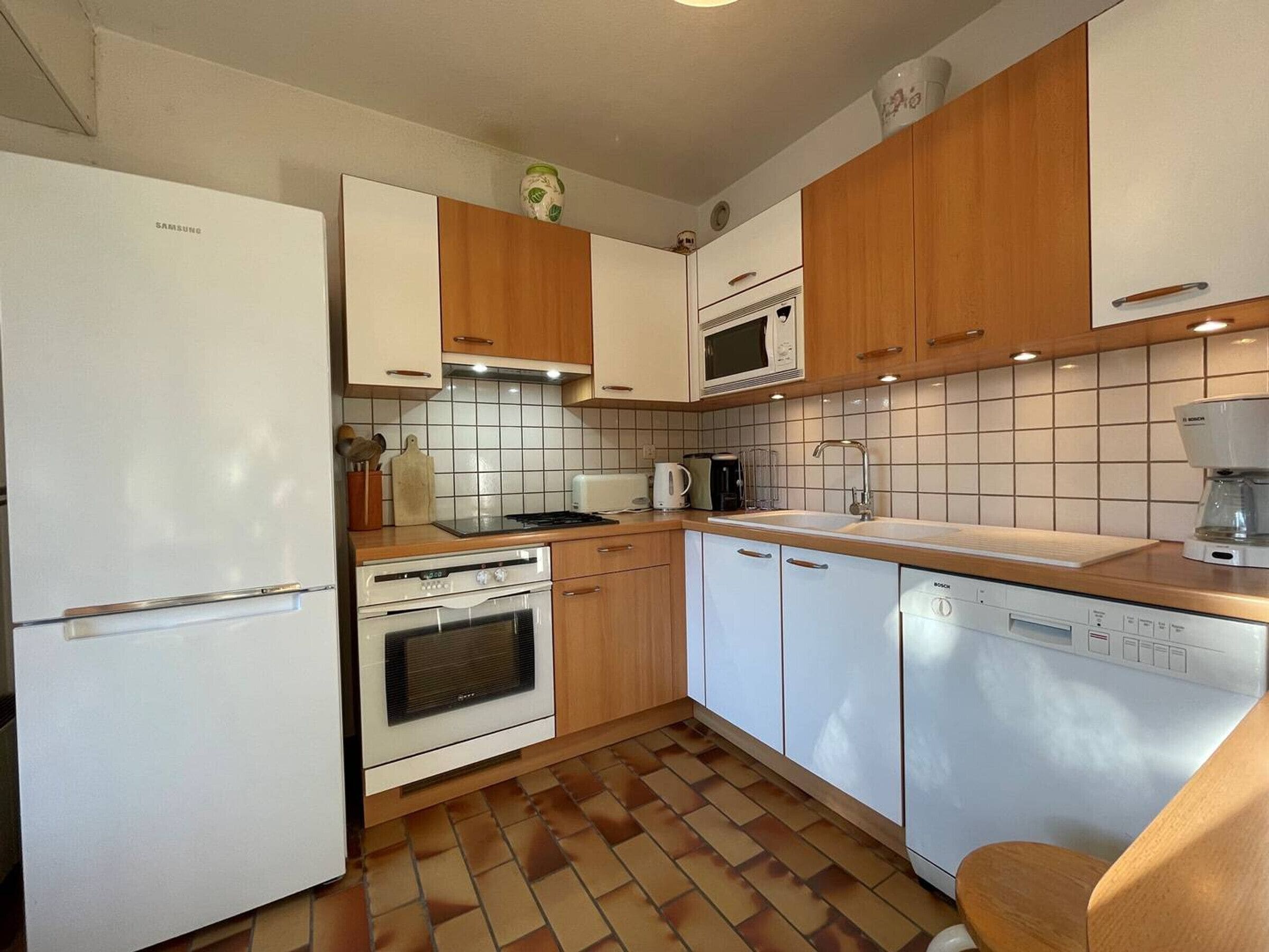 Una cocina funcional con electrodomésticos blancos, armarios de madera y salpicadero de azulejos.