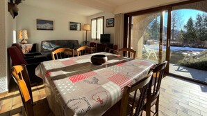 Zona pranzo con tavolo imbandito per sei persone, divano, televisore e vista su un paesaggio innevato attraverso ampie finestre.