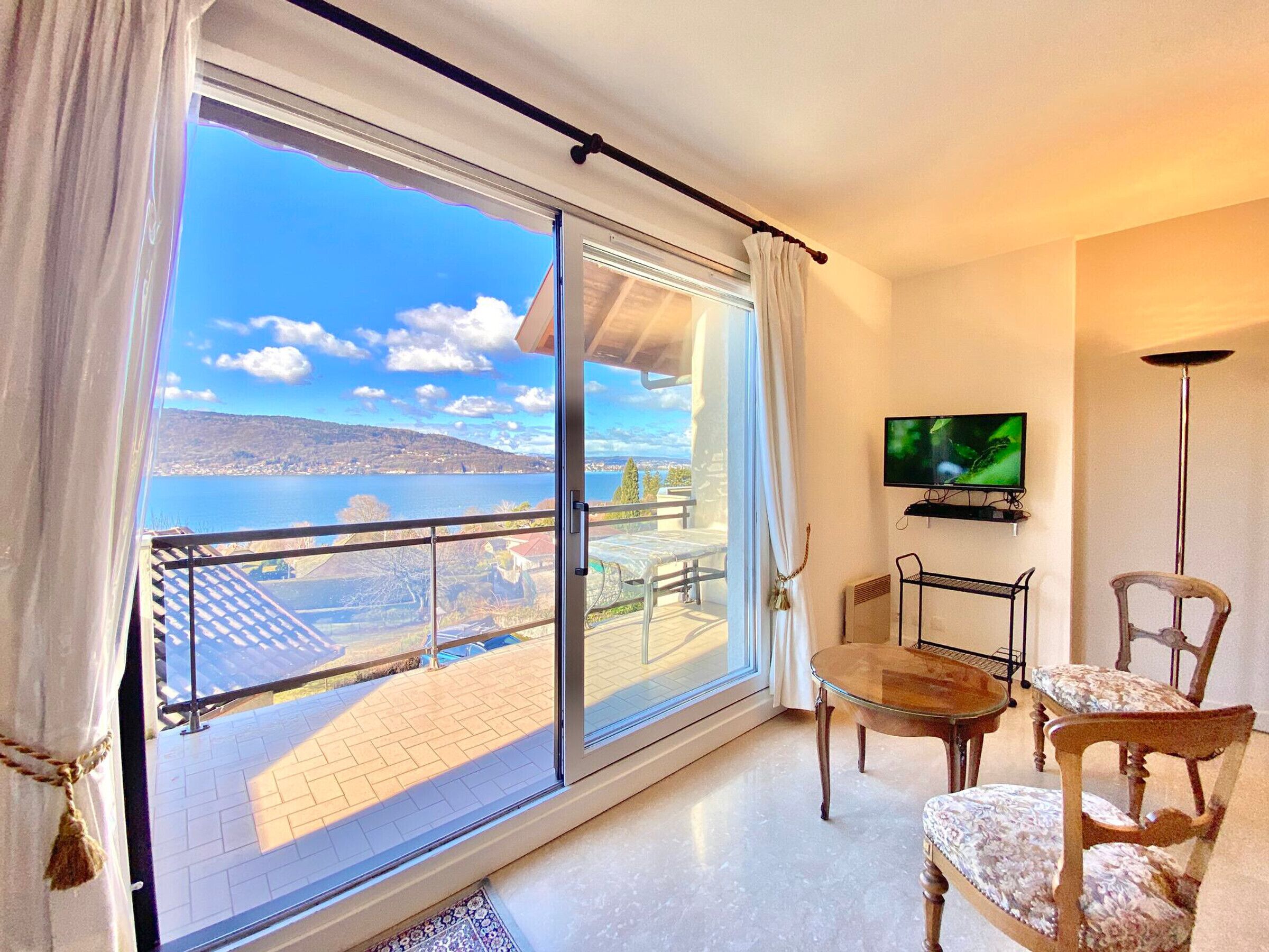 Habitación con un gran ventanal corredero que da a un balcón con vistas panorámicas a un lago y colinas. Se observa un televisor de pantalla plana en la pared, una mesita redonda y dos sillas con tapicería floral.