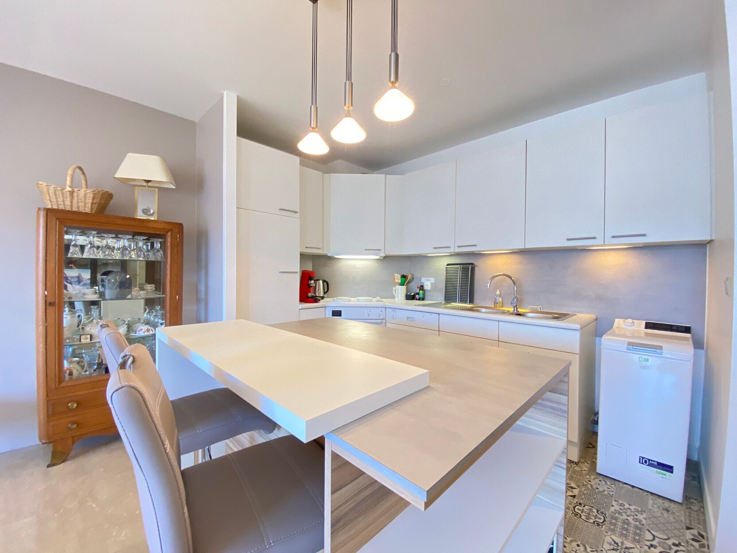 Een moderne keuken met witte kastjes, een centraal eiland, hanglampen en een kleine koelkast.