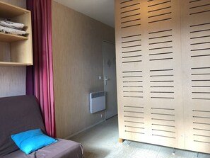 Una habitación con un armario de madera, un sofá gris con un cojín azul y una estantería de madera con objetos doblados.