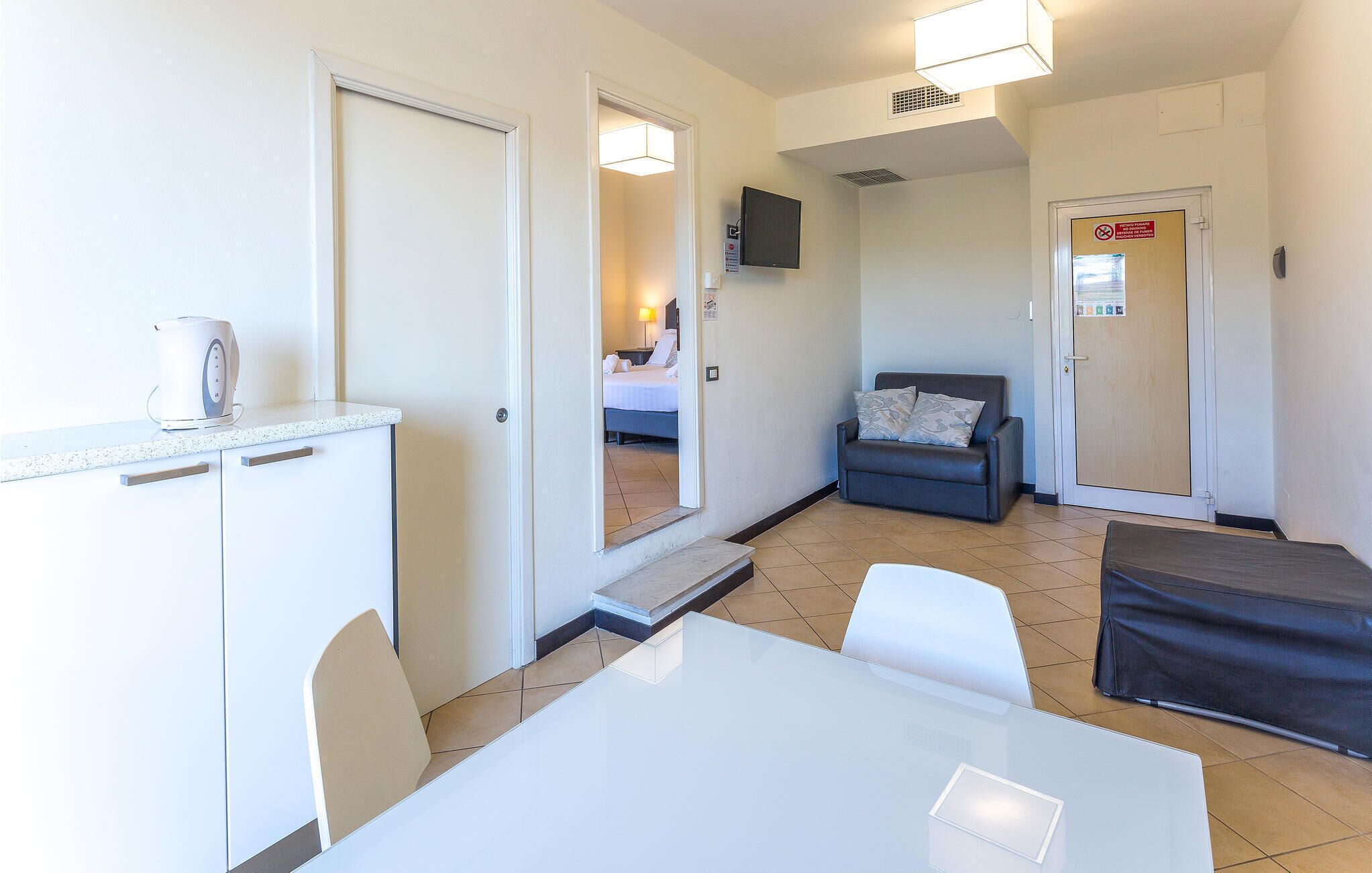 A hotel room with a kitchenette, a dining area, a living area with a sofa, and a bed visible through an open door.