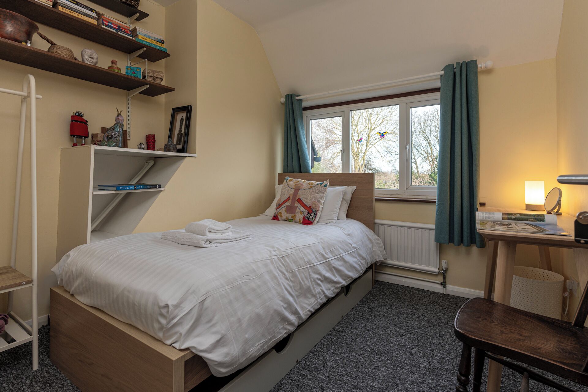 Ein Schlafzimmer mit Bett, Schreibtisch, Stuhl, Heizkörper und Regalen mit Büchern und Dekoration.