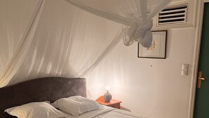 Una cama con ropa de cama blanca y una manta amarilla, una mesita de noche con una lámpara y un respiradero en la pared.
