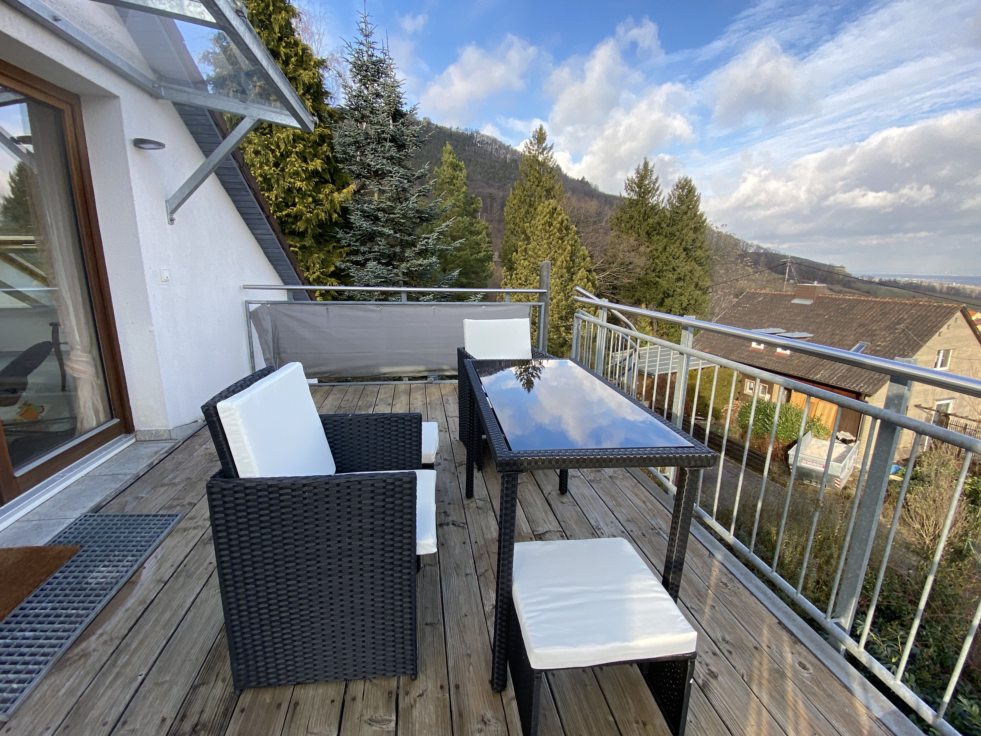 Een balkon met rieten meubels, een glazen deur en uitzicht op een bebost gebied.