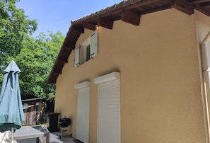 Ein hellbeiges Gebäude mit Holzdach, zwei weißen Fensterläden und einem Grill davor.