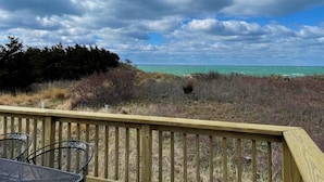 A wooden deck with metal chairs overlooking a beach with clear turquoise water and a cloudy sky.