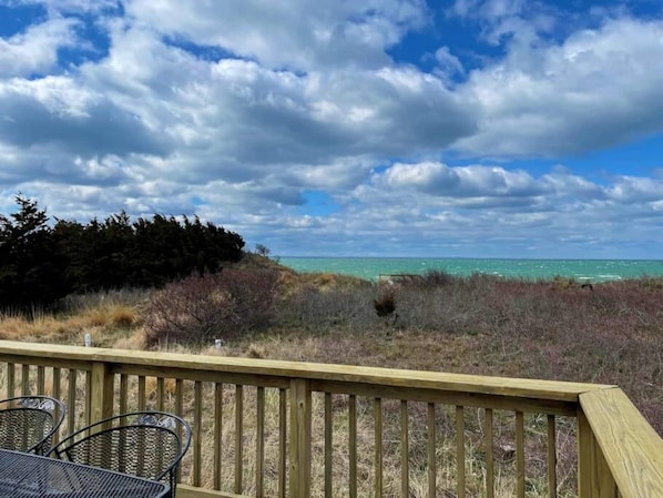 Une terrasse en bois avec des chaises en métal, donnant sur une plage aux eaux turquoise et limpides, sous un ciel nuageux.