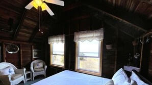 A cozy room with a bed, two chairs, a ceiling fan, and two windows with curtains.