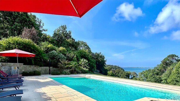 Ein Swimmingpool mit rotem Sonnenschirm, Liegestühlen und Blick auf das Meer.