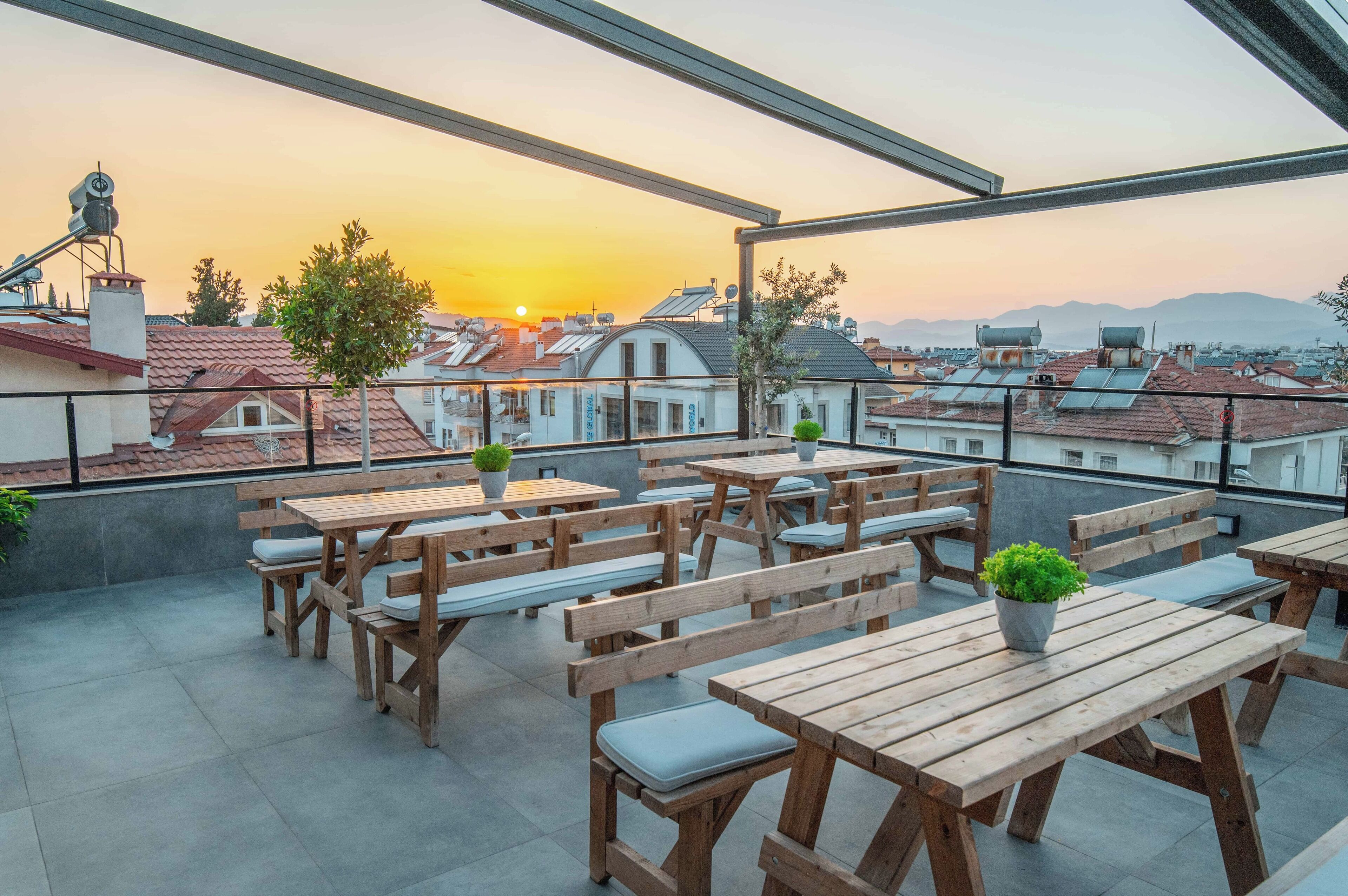 A rooftop terrace with wooden tables and benches, potted plants, and a view of the sunset over a cityscape.