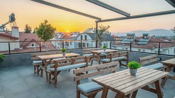 A rooftop terrace with wooden tables and benches, potted plants, and a view of the sunset over a cityscape.