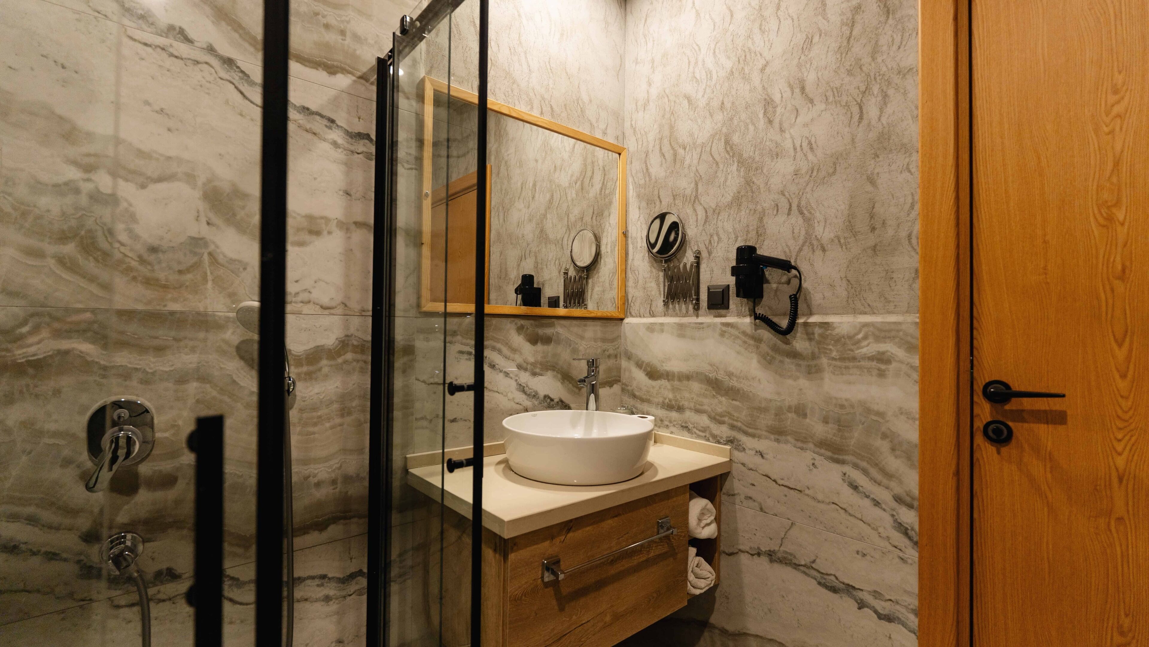 A bathroom with a marble wall, a wooden door, a glass shower enclosure, a sink with a mirror, and a towel rack.